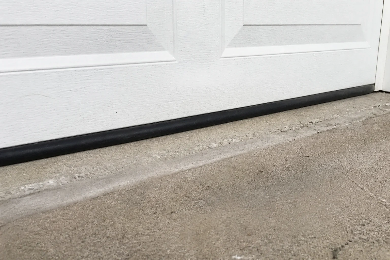 Black bottom seal along a closed white garage door at a concrete threshold.